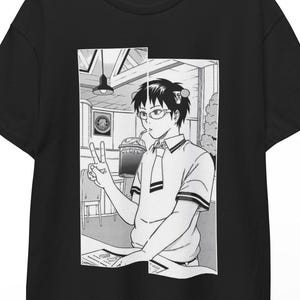 Puede incluir: Camiseta negra con una ilustración de estilo manga. El gráfico muestra a una persona con gafas, haciendo el signo de la paz. La persona está en un café. La camiseta está colgada en una percha de madera.