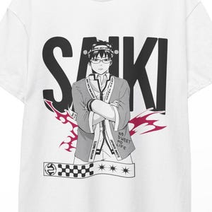 Puede incluir: Camiseta blanca con un gráfico de un personaje de anime y la palabra "SAIKI" en negro. El personaje lleva gafas y un atuendo tradicional. Un diseño de llama roja y negra está debajo.