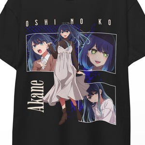 Może przedstawiać: Czarny t-shirt z grafiką z "Oshi no Ko", przedstawiający postać "Akane". Projekt zawiera wiele obrazów postaci z niebieskimi włosami i białą sukienką. Koszulka wisi na drewnianym wieszaku.