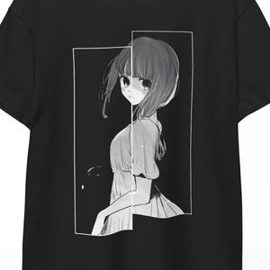 Può includere: T-shirt nero con un'illustrazione in stile manga di una ragazza all'interno di una cornice rettangolare bianca. L'opera è in scala di grigi, con i capelli e l'abito della ragazza in varie tonalità di grigio. La t-shirt è appesa a un appendiabiti di legno.