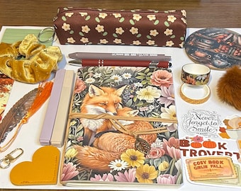 May include: An assortment of fall-themed stationery and accessories. Includes a notebook with a fox illustration, pens, stickers, a scrunchie, a pencil case, a keychain, and decorative tape. The image also features a faux fur pom-pom and a bookmark.