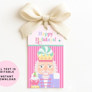May include: A festive gift tag with a cream-colored bow. The tag features a nutcracker illustration with the text "Happy Holidays!". The background is pink with vertical stripes. The nutcracker is holding a dessert.