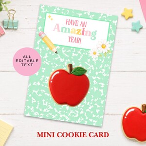 Puede incluir: Una tarjeta rectangular con fondo verde menta moteado que presenta el texto HAVE AN AMAZING YEAR, un lápiz amarillo y dos margaritas blancas. Una galleta roja con forma de manzana y una hoja verde está centrada en la tarjeta.