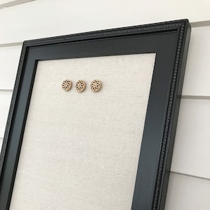 Pode incluir: Um quadro de cortiça preto com uma superfície de tecido de linho. Três alfinetes decorativos de madeira com um design floral estão presos ao quadro.