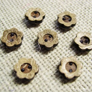 May include: Seven wooden flower-shaped buttons with two black centers. The buttons are arranged in a circle on a beige fabric background.