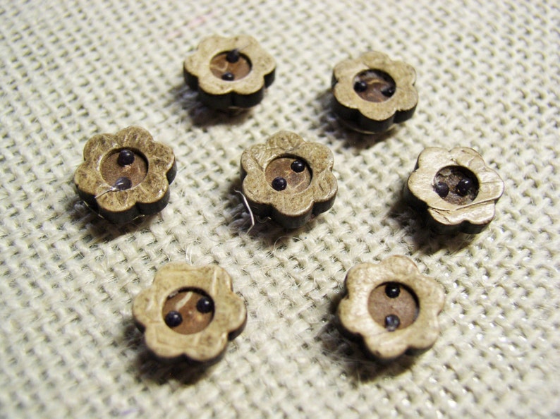 May include: Seven wooden flower-shaped buttons with two black centers. The buttons are arranged in a circle on a beige fabric background.