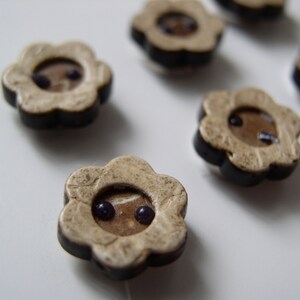 May include: A set of six flower-shaped buttons with a black rim and a beige center. Each button has two small purple dots in the center.