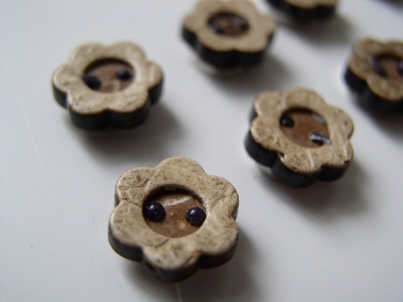 May include: A set of six flower-shaped buttons with a black rim and a beige center. Each button has two small purple dots in the center.