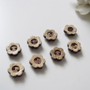 May include: Eight brown wooden flower-shaped buttons with two black centers. The buttons have a light brown, almost gold, color.