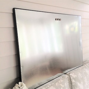 May include: A large, rectangular metal sheet with a black frame. The sheet has a reflective, silver surface and is mounted on a wall. Three small, round fasteners are visible near the top. The item is leaning against a floral patterned cushion.