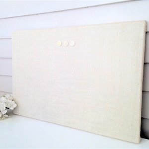 Puede incluir: Un tablón de anuncios rectangular de lino blanquecino con tres pequeños chinchetas redondas de color crema. El tablero está apoyado contra una pared blanca. Un pequeño ramo de flores blancas está delante del tablero. Aproximadamente 60 cm x 40 cm.