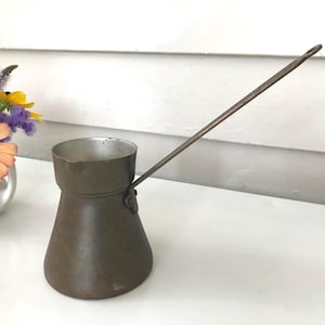 May include: A vintage copper coffee pot with a long, thin handle. The pot has a rounded base and a flared top. The handle extends from the side of the pot. The pot is a muted brown color and appears to be made of metal. A small bouquet of flowers is in the background.