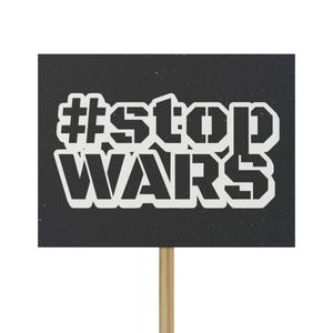 May include: A black sign with the white text "#stop WARS" in a bold, stencil-like font. The sign is mounted on a wooden dowel, suitable for holding up during a protest or demonstration.