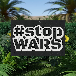May include: Black yard sign with the text "#stop WARS" in white stencil lettering. The sign is displayed outdoors, surrounded by green foliage and a blue sky.
