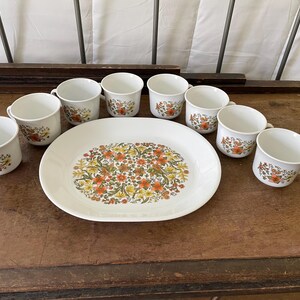 May include: A white ceramic serving platter and seven matching mugs, each decorated with a dense pattern of orange, yellow, and green floral motifs. The platter is oval-shaped, and the mugs feature a simple handle and a smooth, rounded rim.