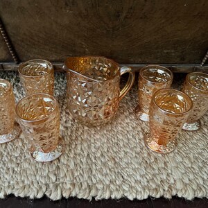 May include: A set of six amber-colored glass tumblers and one matching pitcher, all featuring a raised diamond-cut geometric pattern. The glassware has a reflective, iridescent finish that catches the light, creating a warm, metallic glow.