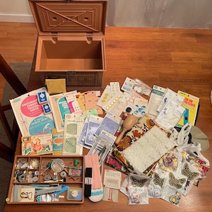 May include: A brown plastic sewing box with an open lid, accompanied by a collection of sewing supplies including packages of buttons, dressmaker carbon tracing paper, needle packs, overall buckles, lace fabric, and various iron-on patches.