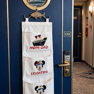 May include: A white fabric hanging organizer with three pockets, suspended from a brass fish-shaped hook on a dark blue door. The pockets feature printed graphics of a cruise ship labeled MOM & DAD, and two cartoon mice labeled LEIGHTON and LANE.