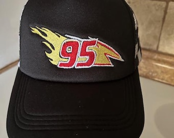 May include: Black trucker hat featuring a flame design with the number 95 in red and yellow. The hat has a black brim and a white mesh back. A fun accessory for fans of racing.