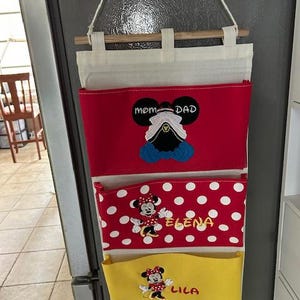 May include: A hanging fabric wall organizer with three pockets. The top pocket is red with a cruise ship icon and the text MOM DAD. The middle pocket is red with white polka dots, a Minnie Mouse graphic, and the name ELENA. The bottom pocket is yellow with a Minnie Mouse graphic and the name LILA.