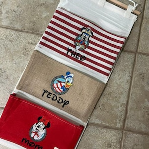 May include: A hanging wall organizer with three fabric pockets. The top pocket is white with red stripes and features Mickey Mouse and the name THEO. The middle pocket is tan burlap with Donald Duck and the name TEDDY. The bottom pocket is red with Minnie Mouse and the name MOM.