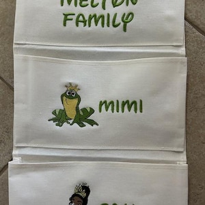 May include: White fabric organizer with three pockets. The top pocket has the text "THE MELTON FAMILY" in green embroidery. The middle pocket features a frog with a crown and the name "MIMI". The bottom pocket shows a princess and the name "CALI".
