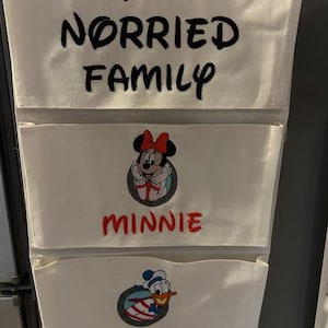 May include: A white fabric hanging wall organizer with three pockets. The top pocket features the black embroidered text THE NORRIED FAMILY. The middle pocket shows an embroidered Minnie Mouse and the red text MINNIE. The bottom pocket displays an embroidered Donald Duck and the black text DONALD.