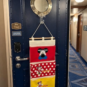 May include: A hanging fabric organizer with three pockets in red, white-polka-dot red, and yellow, featuring embroidered Disney character motifs and the names Mom, Dad, Elena, and Lila, suspended from a hook on a dark blue nautical-style door.
