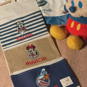 May include: A personalized Disney-themed wall hanging featuring embroidered characters Mickey Mouse, Minnie Mouse, and Donald Duck. The hanging has blue and white stripes, tan and blue sections, and the names "Quinn" and "Marcia" in red.
