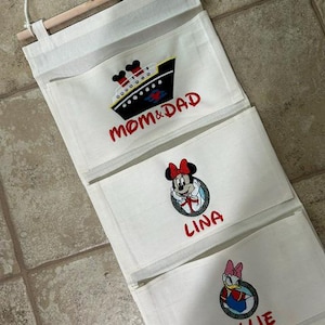 May include: A white fabric wall hanging organizer with three pockets suspended from a wooden dowel. The top pocket features a cruise ship and the text MOM & DAD. The middle pocket shows Minnie Mouse and the name LINA. The bottom pocket features Daisy Duck and the name ELLIE.