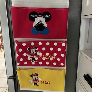 May include: A three-pocket hanging organizer with a cream-colored top and wooden dowel. The top pocket is red with a Mickey Mouse design and the words "Mom Dad". The middle pocket is red with white polka dots and the name "Elena". The bottom pocket is yellow with the name "Lila".