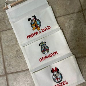 May include: A white fabric wall hanging organizer with three tiered pockets. Each pocket features an embroidered Disney character and red text: Pluto with MOM & DAD, Mickey Mouse with GRAHAM, and Minnie Mouse with HAZEL. It hangs from a wooden dowel.