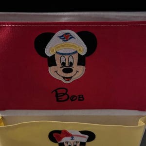 May include: Red and yellow fabric storage pockets with embroidered Mickey and Minnie Mouse faces wearing sailor hats. The name "Bob" is embroidered on the red pocket, and "Kim" is embroidered on the yellow pocket. The word "Family" is embroidered at the top.