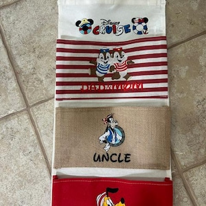 May include: A hanging wall organizer with four fabric pockets. The top pocket features the Disney Cruise logo. The second pocket has red and white stripes with Chip and Dale and the text DAD AND MOM. The third is tan with Goofy and UNCLE. The bottom is red with Pluto and PLUTO.