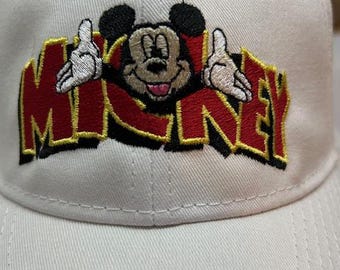 May include: A white baseball cap featuring an embroidered design of Mickey Mouse. The design includes Mickey's face and hands, with the word "MICKEY" in red and yellow lettering. The cap has a curved brim and a structured crown.