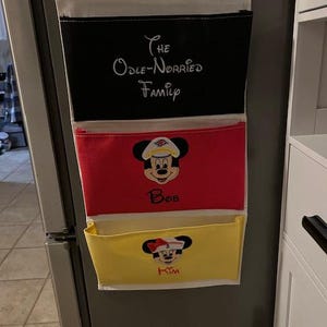 May include: A three-pocket organizer with a white top, black middle, and red and yellow bottom pockets. The black pocket has the text "The Odle-Norried Family". The red pocket has a Mickey Mouse graphic and the name "Bob". The yellow pocket has a Minnie Mouse graphic and the name "Kim".