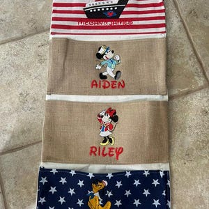 May include: A vertical hanging organizer with four fabric pockets. The top pocket has red and white stripes with a cruise ship and the text Megan & James. Two middle pockets are tan burlap with embroidered characters and the names Aiden and Riley. The bottom pocket is navy blue with white stars, a cartoon character, and the name Emily.