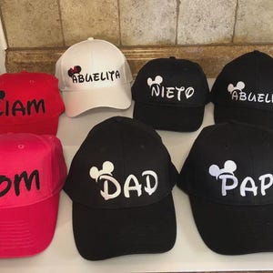 May include: A collection of baseball caps in various colors, including red, pink, white, and black. Each cap features a Mickey Mouse head design and personalized text such as "Liam", "Mom", "Abuelita", "Nieto", "Abuelito", "Dad", and "Papa".