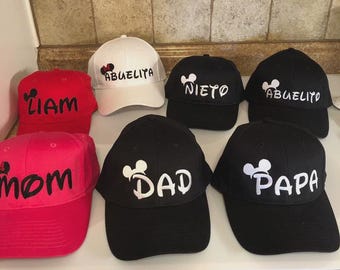 May include: A collection of baseball caps in various colors, including red, pink, white, and black. Each cap features a Mickey Mouse head design and personalized text such as "Liam", "Mom", "Abuelita", "Nieto", "Abuelito", "Dad", and "Papa".