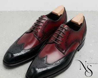 May include: A pair of two-tone leather dress shoes. The shoes are burgundy with black wingtip detailing and brown laces. Shoe trees are inserted. The brand logo "NURSOLES" is in the bottom right corner.