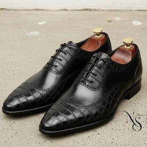 May include: A pair of black leather dress shoes with a textured, crocodile-like pattern on the toe and heel. The shoes have a classic wingtip design with decorative perforations and are laced up. Shoe trees are inserted.