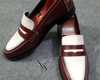 May include: A pair of brown and white leather penny loafers. The shoes have a classic design with a white saddle and white stitching. The shoes are displayed with wooden shoe trees inside.