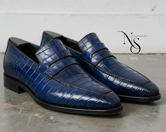 May include: A pair of blue leather loafers with a crocodile skin texture. The shoes have a classic slip-on design with a low heel and a rounded toe. The shoes are displayed on a light gray surface.