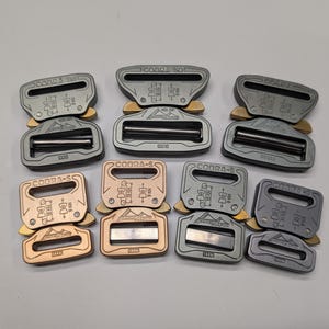 Pode incluir: Uma coleção de fivelas Cobra-S de metal em várias cores, incluindo prata e bronze. Cada fivela tem um design retangular com uma barra central e o texto "COBRA-S". As fivelas estão dispostas sobre uma superfície branca.