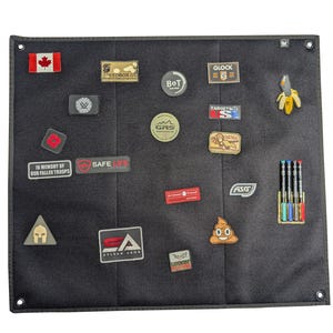 Può includere: Una bacheca in feltro grigio scuro con varie toppe e spille attaccate. Le toppe includono una bandiera canadese, il testo "SAFE LIFE" e il testo "IN MEMORY OF OUR FALLEN TROOPS". Altre toppe hanno il testo "GLOCK", "TARGETS" e "ASG". Sono attaccate anche una spilla a banana e una spilla emoji a cacca.