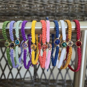 Puede incluir: Una colección de pulseras de macramé coloridas con detalles de piedras preciosas. Las pulseras, en tonos de verde, morado, amarillo, rosa, azul, blanco y rojo, se exhiben en una barra de metal. Cada pulsera tiene una piedra preciosa central.