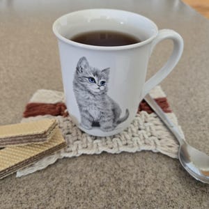 May include: A white ceramic mug filled with dark liquid, featuring a gray kitten illustration. The mug sits on a rectangular macrame coaster with cream and rust-colored yarn. Two rectangular chocolate wafer cookies and a silver spoon are nearby.