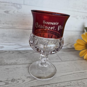 Puede incluir: Una copa de recuerdo vintage con la parte superior de color rojo rubí y una base transparente estampada. La copa está grabada con "Souvenir Coalport, Pa." en letras plateadas. El tallo es transparente y la copa está sobre un fondo de madera clara.
