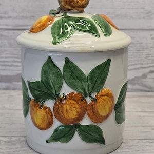 May include: A white ceramic canister with a matching lid featuring a three-dimensional lemon handle. The body and lid are decorated with hand-painted orange citrus fruits and green leaves, creating a vibrant botanical pattern on the smooth surface.