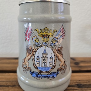以下が含まれることがあります： 金属製の蓋が付いたライトグレーの陶器製ビアジョッキ。正面には、城のイラストを挟んで2頭のライオンが描かれたカラフルな紋章があり、その上には王冠と旗が飾られています。下部の青いバナーには「København」という文字が記されています。